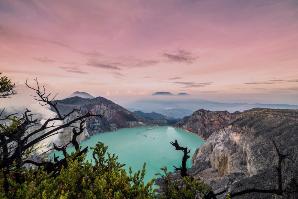 A turquoise volcanic crater lake surrounded by rocky cliffs under a soft pink sky